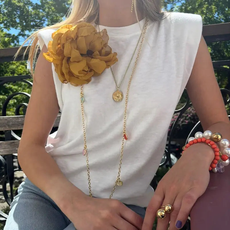 Frau mit goldenem Layering-Schmuck: langer Gliederkette mit bunten Anhängern, Münzanhänger-Halskette, Perlen- und Korallarmband sowie senfgelbem Stoffblumen-Brosche